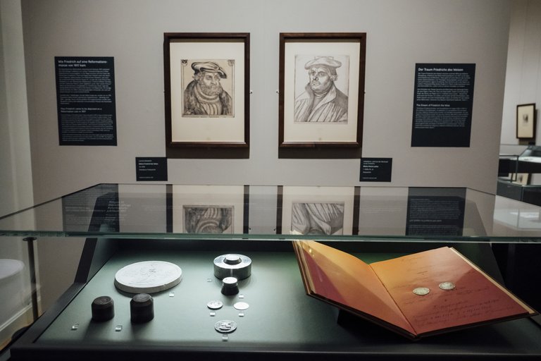 Vitrine mit einem Buch und Münzen, dahinter zwei Bilder an der Wand