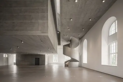Ein weitläufiger, grauer Raum mit Betondecke und einer geschwungenen Treppe.