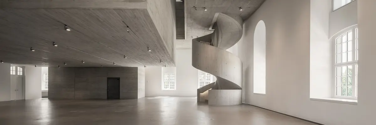 Ein weitläufiger, grauer Raum mit Betondecke und einer geschwungenen Treppe.