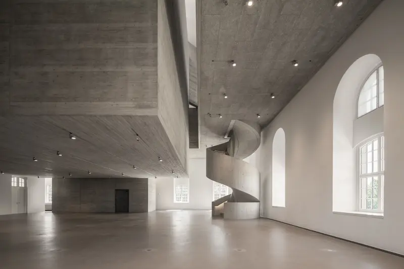 Ein weitläufiger, grauer Raum mit Betondecke und einer geschwungenen Treppe.
