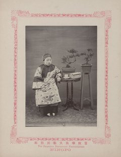 Person im Gewand und mit Kopfbedeckung sitzt neben Tischchen mit Geschirr und Bonsais