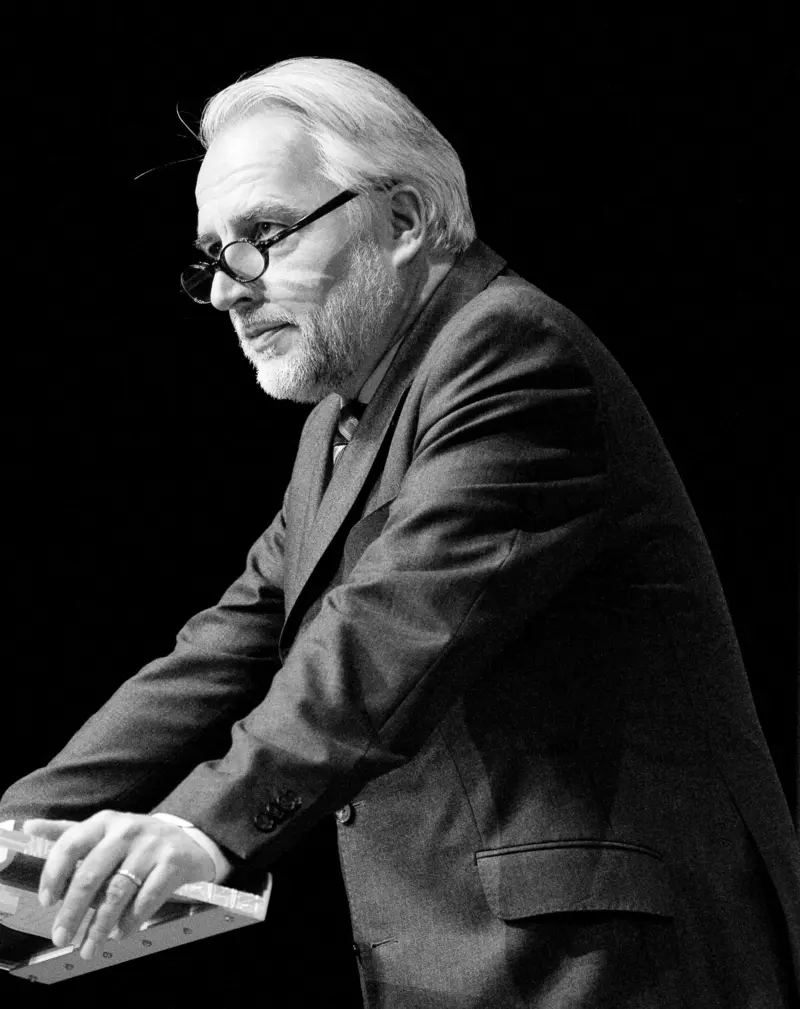 Seintenansicht eines Mannes in Schwarzweiß, der an einem Stehpult lehnt und ernst schaut