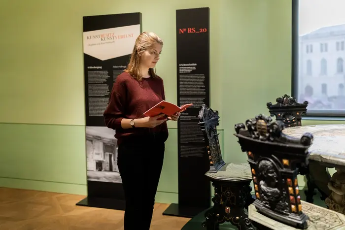 Besucherin in einer Ausstellung vor einem historischen Tisch mit Stühlen 